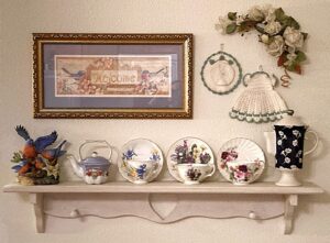 A small decorative wall shelf has three teacup and saucer sets on display next to a tiny china teapot and bluebird family figurine under a framed print of a bluebird pair with the word Welcome between them. A small upright lidded hot chocolate pot is set on the opposite side of the shelf from the bluebird family, and above it is a small faux floral swag attached above a set of a vintage crocheted hat and dress pot holders, too pretty not to display. Welcome into the dining area of my home.